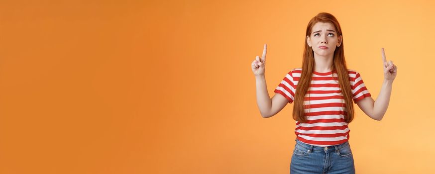 Hesitant upset cute redhead woman weighing choices look doubtful uncertain, frowning thoughtful, ponder decision, look pointing up unsure, have doubts, stand insecure orange background.