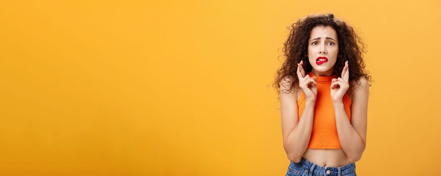 Worried and anxious silly caucasian female. with curly hair in red lipstick and cropped top biting lower lip nervously looking concerned crossing fingers for good luck making wish over orange wall.