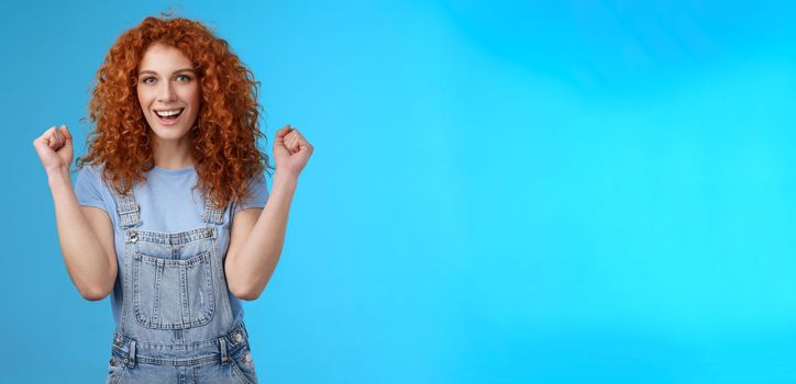 Hooray successful day. Cheerful chrismastic redhead girl summer dungarees clench fists joyfully smiling broadly determined achieve success, triumphing winning game standing upbeat blue background.