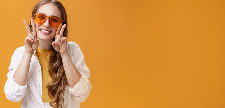 Stylish enthusiastic and charismatic young party girl in trendy sunglasses and white blouse over t-shirt showing peace gestures near face and smiling cute at camera against orange background. Copy space