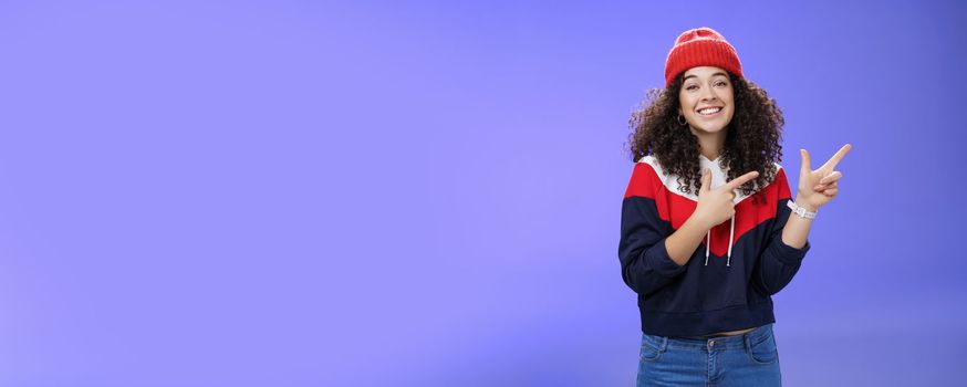 Cute female friend pointing at upper right corner with finger pistols as smiling broadly at camera wearing warm winter hat and sweatshirt skiing over blue background joyfully. Emotions, weather and fashion concept