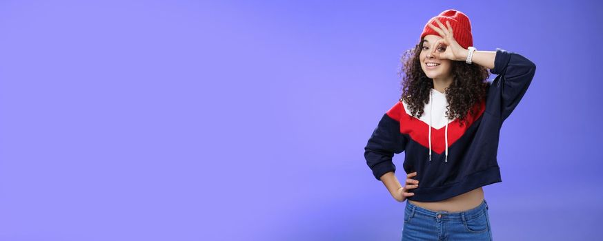 Okay girl agrees. Friendly-looking happy and charming young curly-haired girl in warm beanie showing ok gesture or circle over eye as peeking at camera and smiling broadly with joy over blue wall.