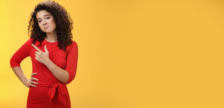 Upset disappointed and unimpressed attractive young european curly-haired woman in red dress pursing lips in gloomy face, frowning displeased as pointing at upper left corner from regret and dislike.