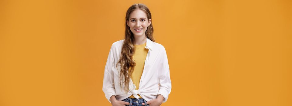 Lifestyle. Stylish young female designer wants help friend shopping holding hand in pockets smiling joyfully and self-assured at camera wearing trendy blouse over yellow t-shirt posing over orange wall.