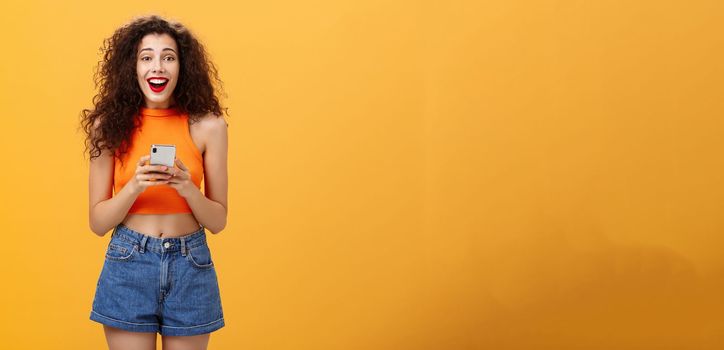 Indoor shot of surprised happy and delighted attractive european female with curly haircut in cropped top holding smartphone gazing at camera amazed and pleased receiving awesome news via messages. Technology concept