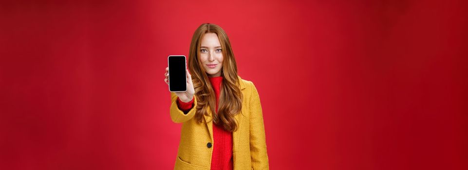 Attractive and stylish ginger girl with confident. glance and self-assured smile pulling mobile phone at camera showing smartphone screen grinning delighted as posing against red background.