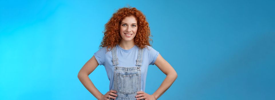 Attractive sassy motivated redhead european female curly-haired hold hands waist empowered express confidence self-assured look forward ready win conqueror, stand blue background. Copy space