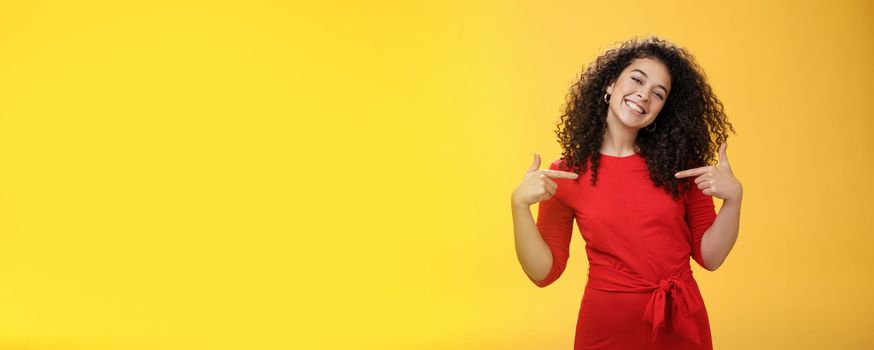 Lifestyle. Proud and satisfied ambitious successful female student in red dress standing pleased smiling and pointing at herself as if bragging about own achievements happily and glad over yellow wall.