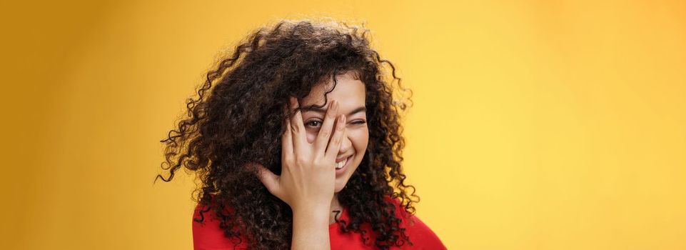 Lifestyle. Close-up shot of sensual and playful attractive girlfriend with curly hair covering face with palm and peeking through fingers with happy tender smile anticipating surprise over yellow background.