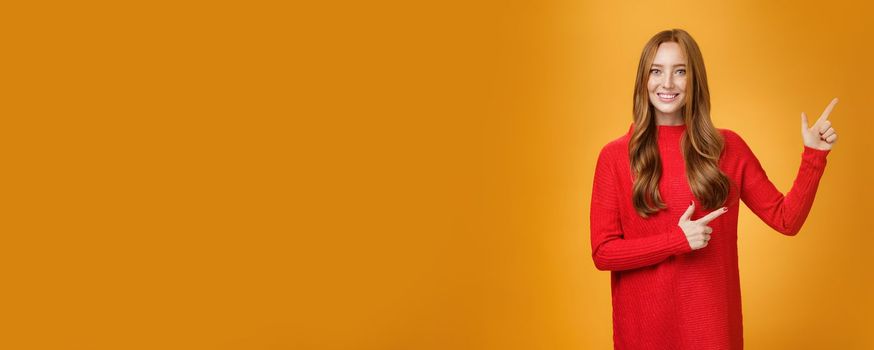 Lifestyle. Friendly-looking joyfuly and energized redhead female in red sweater pointing at upper right corner promoting advertisement with broad delighted and tender smile posing against orange background.