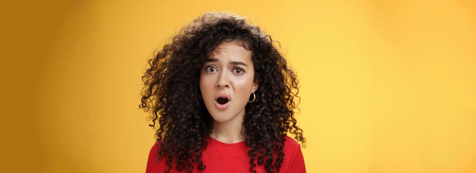 Lifestyle. Confused and frustrated young questioned woman with curly hair open mouth and raising eyebrow in surprise being displeased with unfair situation standing clueless and upset over yellow background.