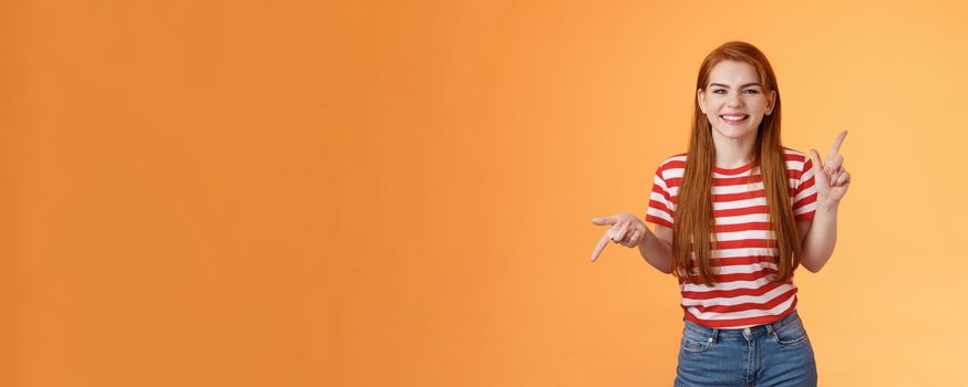 Carefree daring redhead cheerful girl dancing, pointing sideways, showing different sides, left right copy space, laughing and smiling broadly, joyfully recommend cool product, orange background.