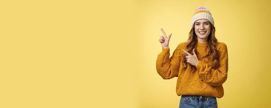 Charming carefree smiling young 20s woman wearing hat sweater pointing upper left corner sideways sharing interesting link awesome promotion delighted recommend cool product, yellow background.
