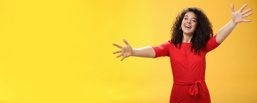 Come into my arms. Portrait of friendly and loving, caring charming woman with curly hair in red casual dress spread hands as wanting give hug smiling broadly at camera giving warm welcome or cuddle.