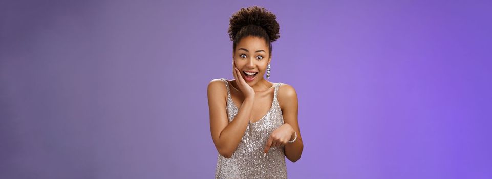 Attractive surprised charismatic african-american woman in silver glittering stylish dress widen eyes gasping amazed touch cheek impressed astonishing perfect gift pointing down cannot believe.