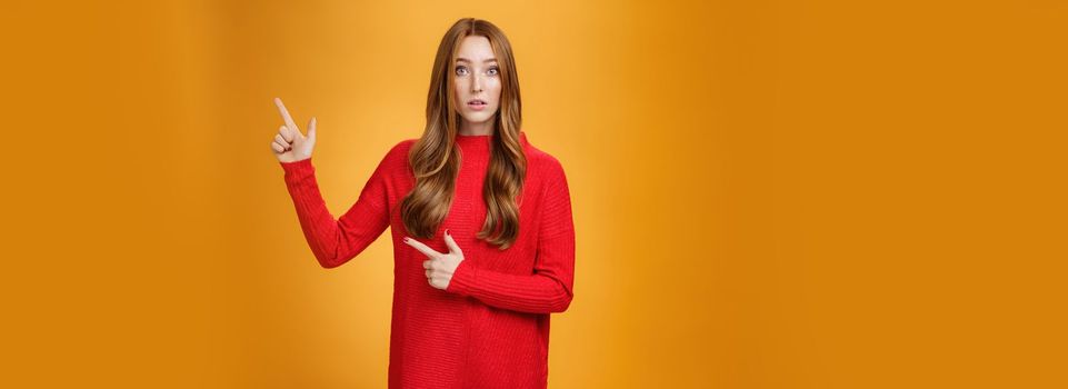 Portrait of elegant and stylish redhead 20s woman with freckles and long hair in knitted red dress pointing at upper right corner looking questioned and impressed, speechless over orange background.