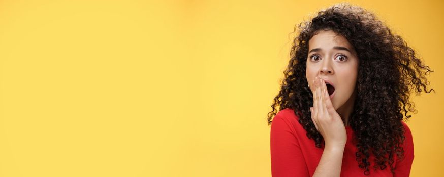 Close-up shot of concerned and shocked worried woman with curly hairstyle open mouth wide and covering it with palm popping eyes at camera impressed and upset over yellow background. Emotions and faciale xpressions concept