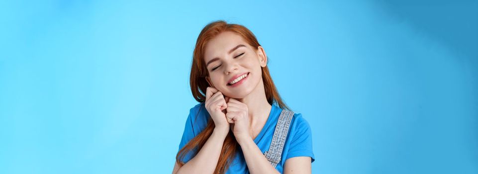 Glad cute tender redhead female, tilt head dreamy lean palms, close eyes and smiling lovely, daydreaming, recall nice memories, cherish moments, stand silly blue background nostalgic mood.