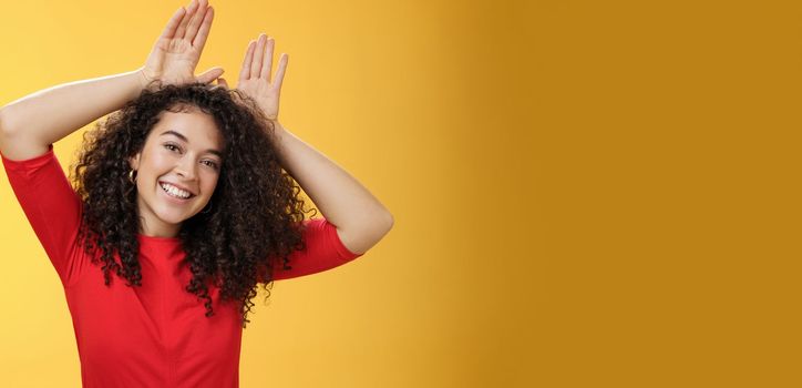 Close-up shot of charismatic playful and tender young kind woman with curly hair playing having fun showing bunny ears with hands on head tilting head joyfully fooling around and smiling at camera. Copy space