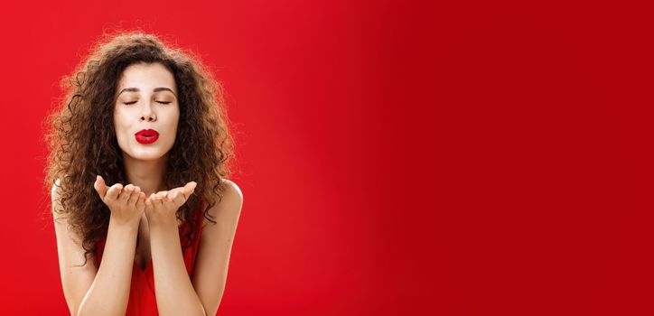 Tender and gentle stylish caucasian girl with curly hairstyle. and red lipstick bending towards camera with slight smile closed eyes and palms near folded lips blowing kiss at camera romantically.