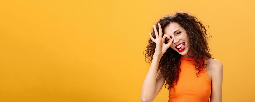 Carefree optimistic and energized good-looking woman. with curly hairstyle tilting head and sticking out tongue playfully showing okay or excellent sign over eye posing near orange background.