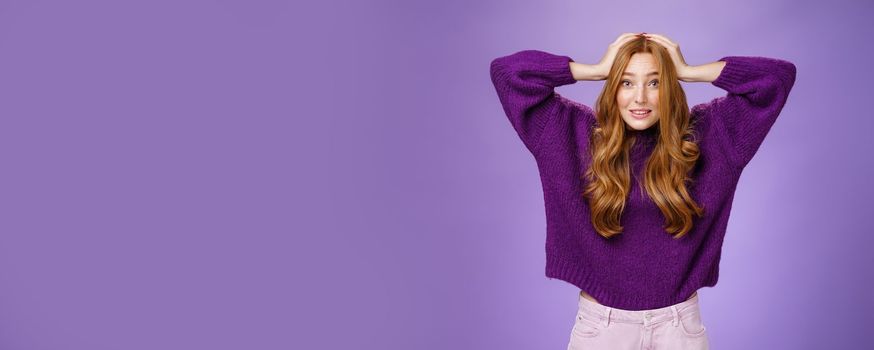 Cute european redhead 20s woman acting nervous, holding hands on head raising eyebrows in worry and anxiety, panicking being troubled think up plan posing over purple background. Copy space
