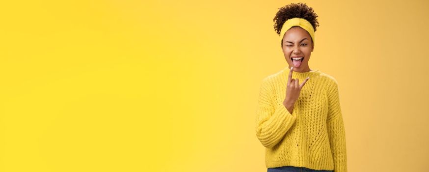 Excited charming smiling daring sassy young african american woman having fun show tongue rock-n-roll heavy metal gesture winking happily enjoying cool awesome party, standing yellow background.