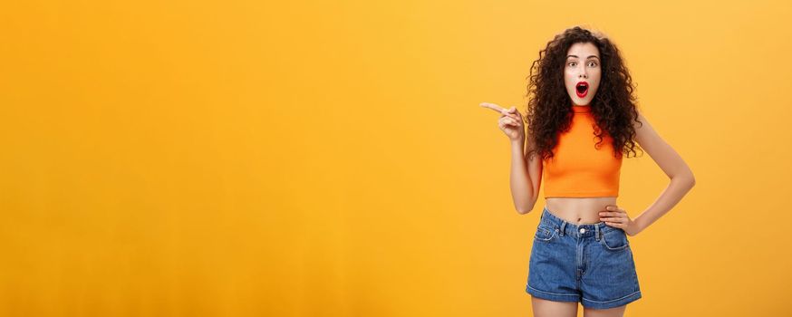 Silly and moody european party girl with curly hairstyle. in red lipstic and stylish orange cropped top complaining on bad weather raising hands pointing and looking up with displeased unhappy face.