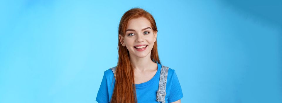 Friendly confident pleasant redhead woman helpfully look camera, smiling joyfully, have conversation customer, give advice, stand upbeat good positive mood blue background, grin amused.
