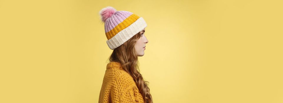 Profile shot attractive female curly hairstyle wearing hat knitted sweater standing queue order hot warm drink look left normal casual expression, posing yellow background studio. Copy space