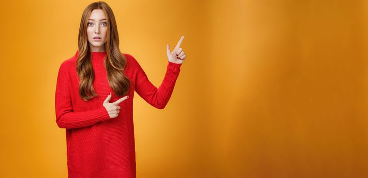 Portrait of elegant and stylish redhead 20s woman with freckles and long hair in knitted red dress pointing at upper right corner looking questioned and impressed, speechless over orange background.