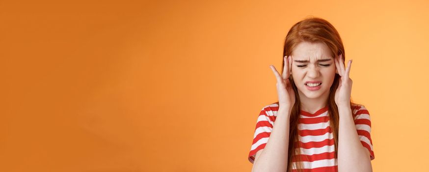 Dizzy uneasy cute redhead female student suffering huge headache, cringe, frowning clench teeth painful feeling, touch temples, feel pain head, awful migraine, asking painkillers, orange background.