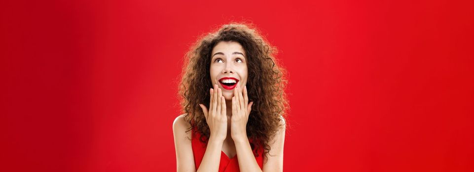 Waist-up shot of amused and entertained happy young dreamy girlfriend. in stylish dress holding palms on chin and opening mouth with broad smile looking delighted up as if seeing miracle over red wall.