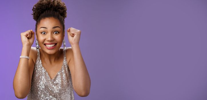 Excited african woman in elegant silver dress brilliants smiling thrilled winning clench raised fists grinning triumphing rejoicing receive perfect news celebrating, standing blue background.