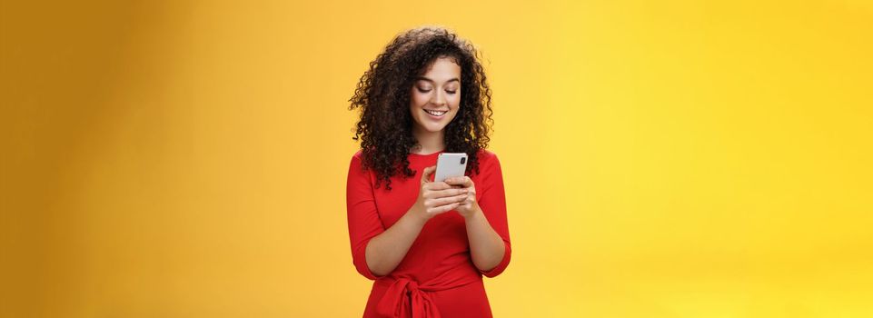 Lifestyle. Gil sending message spread news across social network having party inviting friends via smartphone holding mobile phone in hands smiling broadly at device screen as posing over yellow background.