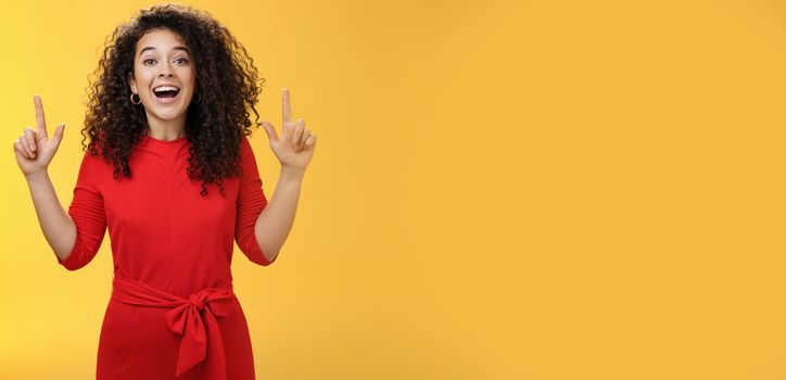 Enthusiastic happy curly-haired young european woman feeling happy present awesome copy space, raising hands pointing up and smiling with delight and admiration over yellow background.