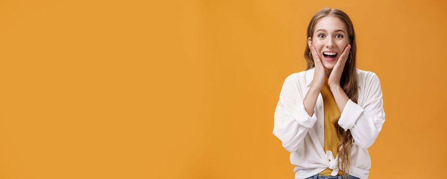 Excited and amused pretty young stylish woman with tattoo in trendy outfit touching cheeks from surprised and thrill gazing entertained at camera hearing awesome news, posing over orange wall. Body language concept