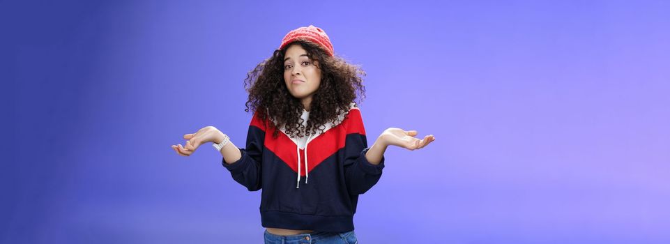 Upset girl cannot get clue what happened shrugging turning away as pouting feeling questioned and unsure what answer, standing clueless and puzzled in sweatshirt and stylish hat over blue background.