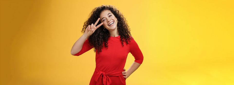 Waist-up shot of friendly tender and feminine cute woman with curly hairstyle tilting head and smiling, laughing and showing victory or peace gesture at camera over yellow background.