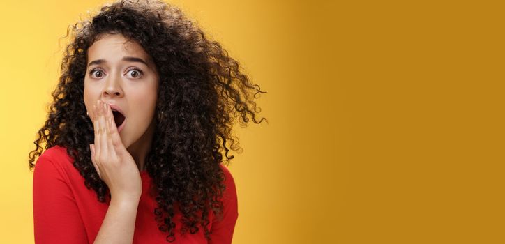 Close-up shot of concerned and shocked worried woman with curly hairstyle open mouth wide and covering it with palm popping eyes at camera impressed and upset over yellow background. Emotions and faciale xpressions concept