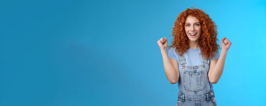 Hooray successful day. Cheerful chrismastic redhead girl summer dungarees clench fists joyfully smiling broadly determined achieve success, triumphing winning game standing upbeat blue background.