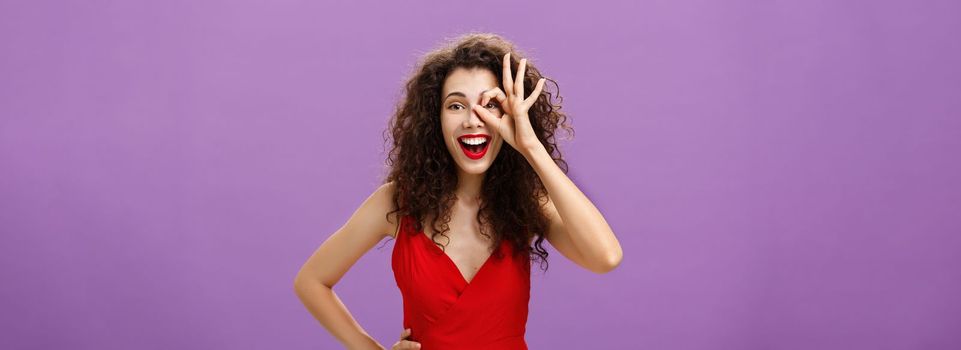 I got zero worries. Portrait of joyful friendly and happy charming woman with curly hairstyle in elegant red dress holding hand on waist smiling amused showing okay sign over eye looking through it.