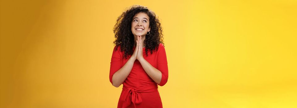 Lifestyle. Dreamy excited cute b-day girl with curly hair in cute red dress rubbing palms together near chest as hands in pray smiling looking up delighted and hopeful making wish over yellow background.