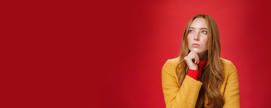 Close-up shot of determined and focused creative thoughtful redhead female looking at upper left corner touching chin, thinking, making choice or remembering information over red background.