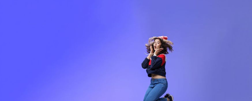 Lifestyle. Surprised and amazed amused good-looking woman with curly hair in winter beanie and sweatshirt open mouth from joy and amazement jumping over blue background having fun feeling playful.