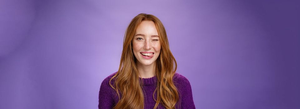 Lifestyle. Close-up shot of sincere and happy funny cute redhead female with long ginger hair winking happily and sticking out tongue childish and carefree posing cheerful over purple background.