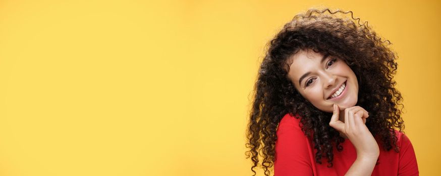 Close-up shot of charming flirty and silly curly-haired young woman making faces as trying get what wants, smiling tilting head on shoulder and touching lip standing cute over yellow background. Romance, people and beauty concept