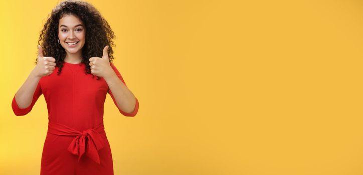 Excellent idea, nice job. Portrait of supportive delighted and happy charming female friend showing thumbs up as standing in red dress over yellow wall smiling giving positive reply, liking concept.