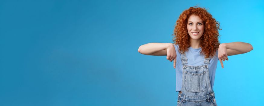 Lifestyle. Charismatic sassy flirty redhead daring ginger girl curly haircut pointing down index fingers smiling broadly enthusiastic explore new store pointing promo like cool advertisement blue background.