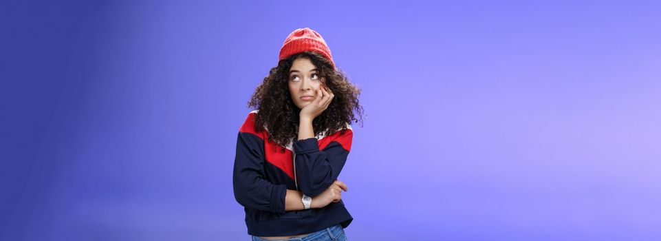Portrait of frustrated and bored lonely girl with curly hair in winter hat holding hand across chest leaning head on palm, looking silly and gloomy at upper right corner, thinking feeling boredom.